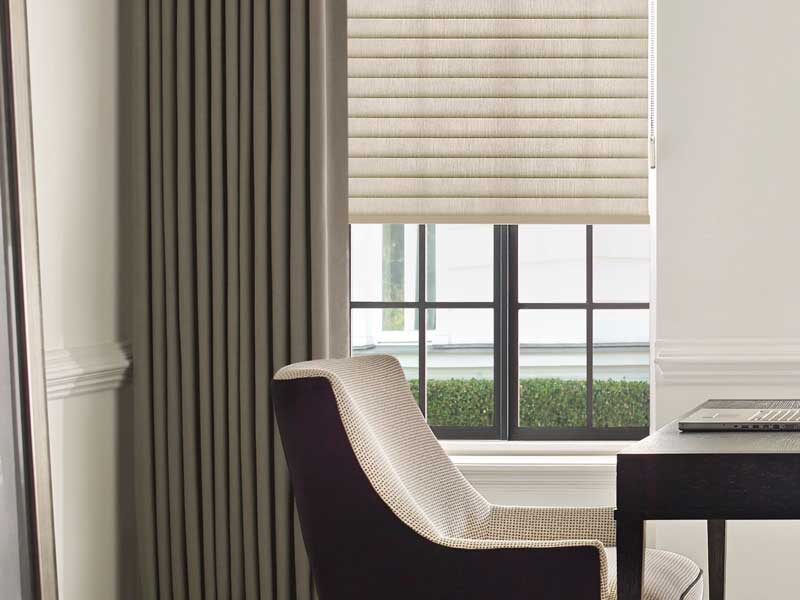Sonnette® Cellular Roller Shades with Custom Drapes. A closeup of a desk with a laptop on it and a chair. The drapery is visible from the side as the bold color pops against the neutral tones in the room.